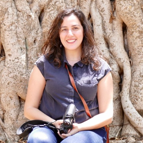 Beatriz is sitting on the roots of big trees in Gondar, Ethiopia, holding a pair of binoculars