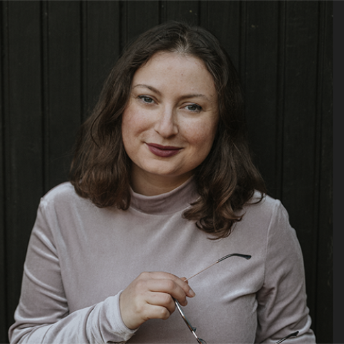 Milena hält eine Brille in der Hand und steht vor einer schwarzen Holzwand