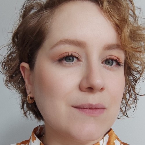 Das Bild zeigt eine Frau mit kurzen, lockigen, blonden Haaren. Sie trägt ein orangefarbenes Oberteil mit einem weißen, unregelmäßigen Muster und goldene Ohrringe. Ihr Make-up ist dezent, mit natürlichen Tönen und leicht betonten Wimpern. Der Hintergrund ist neutral und schlicht.