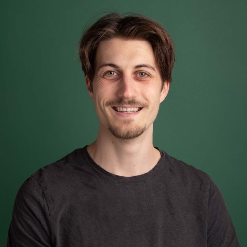 A man smiling in front of a green photo studio background. Brown hair and grey T-shirt.