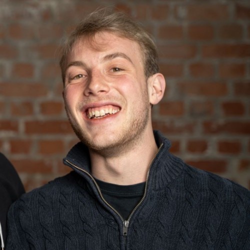 Stefan Wilhelm (ein blonder Mann Mitte 20) steht lächelnd vor einer Backsteinwand. Er trägt einen dunkelblauen Pullover.