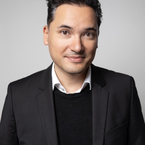 Ein Mann in schwarzem Blazer steht vor einem grauen Hintergrund und lächelt.