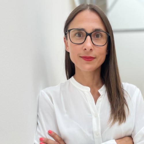 SuSa lehnt an einer weißen Wand in der Agentur. Sie hat eine weiße Bluse an. Glatte Haare und eine Brille auf.