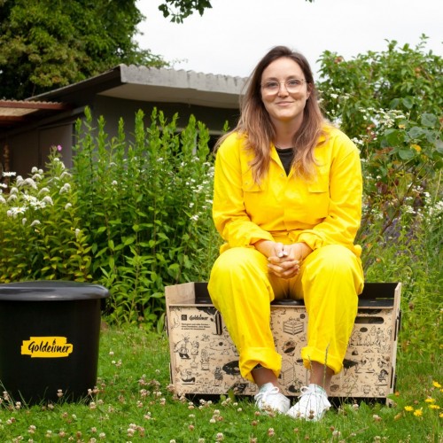 Tanja sitzt im gelben Overall auf einer Trockentoilette von Goldeimer und lächelt in die Kamera. Im Hintergrund sind blühende Sträucher.
