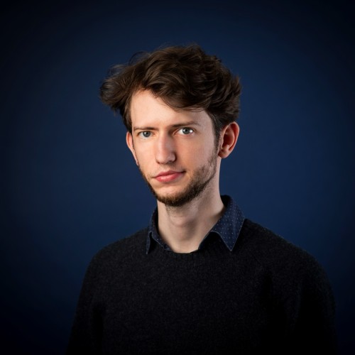 Ein junger Mann Ende 20 vor einem dunkelblauen Fotohintergrund. Er hat braune Haare und leichen Bartwuchs, sowie einen schwarzen Pullover an. Er lächelt leicht in die Kamera. 