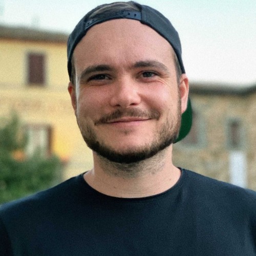 Junger Mann, leicht lächelnd, mit schwarzem Shirt und unscharfen Häusern im Hintergrund.