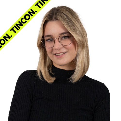 Junge Frau mit schulterlangen, blonden Haaren und Brille, trägt einen schwarzen Rollkragenpullover und lächelt