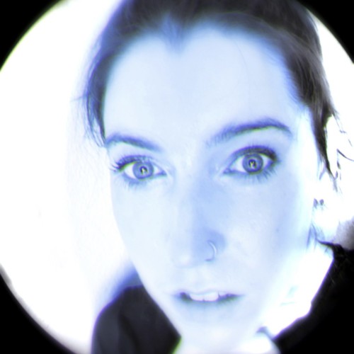 Photograph of the artist Lyndsey Walsh, cropped in a circular shape with a blue tint. Lyndsey has dark brown hair and pale white skin. The artist is looking directly at the camera.