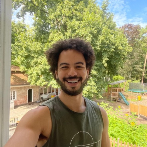Auf dem Foto ist ein Mann mit lockigem Haar und heller Haut vor einem Garten zu sehen.