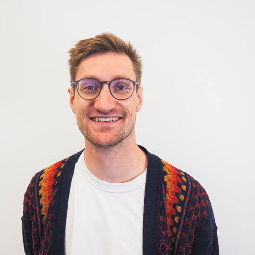 Ein junger Mann mit Brille und Drei-Tage-Bart lächelt in die Kamera. Er trägt eine blaue Wolljacke mit roten Streifen und steht vor einer weißen Wand.