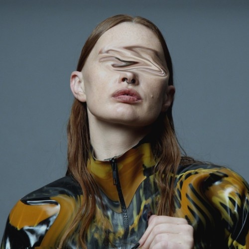A portrait with shoulders, of a person with light skin and red hair in a tree green latex shirt with wavy orange and yellow stripes. On a grey studio background and with digitally edited wavede eyes. 