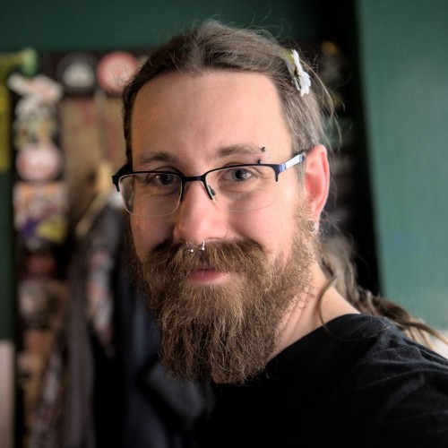 Portrait einer weißen Person mit Bart und langen Braunen Haaren. Er trägt eine Brille, hat einen Nasenring und einen Augenbrauenpiercing, sowie eine weiße Blume im Haar.