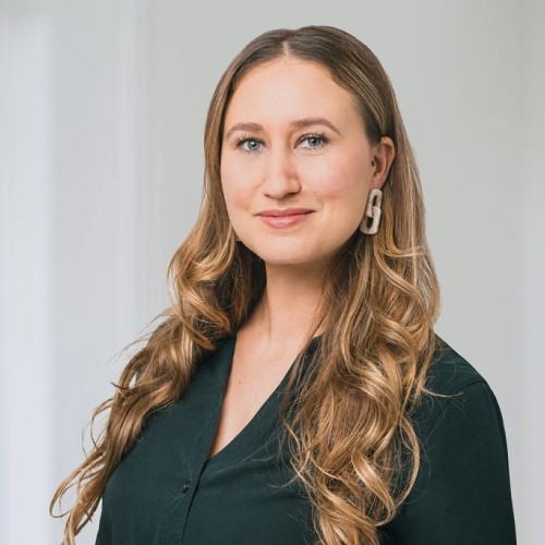 Das Foto zeigt eine Frau mit langen Haaren, die zur Kamera blickt. Sie trägt eine dunkle Bluse und steht vor einem hellen Hintergrund.
