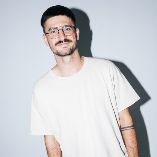 Auf dem Bild Alex Elsner zu sehen. Er trägt ein weißes T-Shirt, eine Brille it dünnem Gestell, hat braune Haare und braunen Bart.