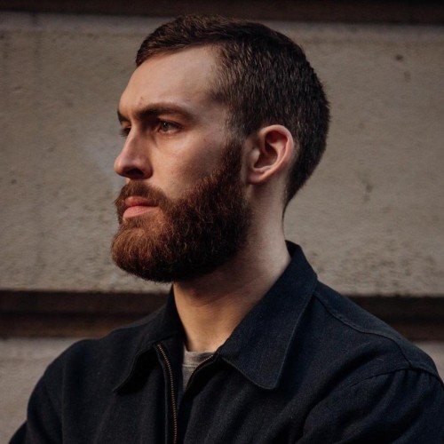 A white man with a red beard looks to the left of the camera.