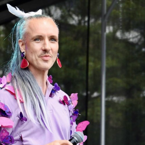 Person mit langen, glatten grauen Haaren, einem Schnauzbart, lilafarbenen Ohringen, einem lilafarbenen, mit Schmetterlingen benähten Oberteil und einem weißen Vogel auf dem Kopf