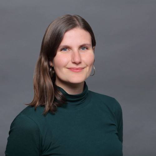 Man sieht eine weiblich gelesene Person im Portraitformat vor grauem Hintergrund. Sie trägt einen petrolfarbenes Langarmshirt und goldene Kreolen. Sie lächelt.   