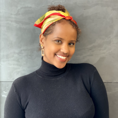 A photo of Arda Awais - A black woman wearing a black turtle neck and a yellow with red tones headband. Her hair is curly brown tied up as a top knot. 