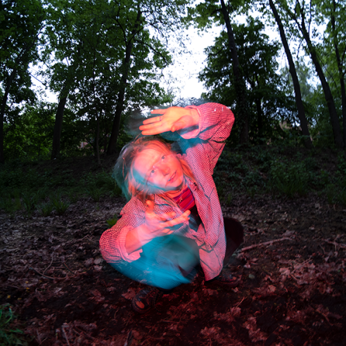 Pink lit image of a white female, slightly blurry in motion, against a dark forest backdrop. 