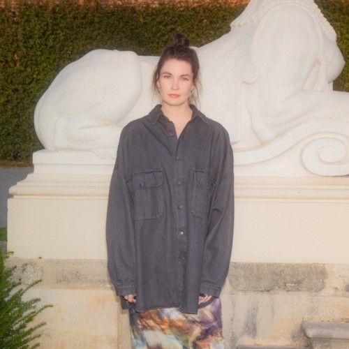 Annekathrin Kohout, eine junge Frau mit heller Haut und dunklen Haaren, steht vor einer weißen Steinskulptur, die eine Sphinx darstellt. Sie trägt ein oversized schwarzes Hemd, das locker über einen fließenden Rock fällt. Der Rock hat ein buntes Muster in violett, orange und blauen Tönen, das an ein Gemälde erinnert. Ihre Haare sind zu einem lässigen Dutt gebunden, und sie blickt mit einem ruhigen, neutralen Gesichtsausdruck in die Kamera. Die Szene ist von warmem Sonnenlicht beleuchtet.