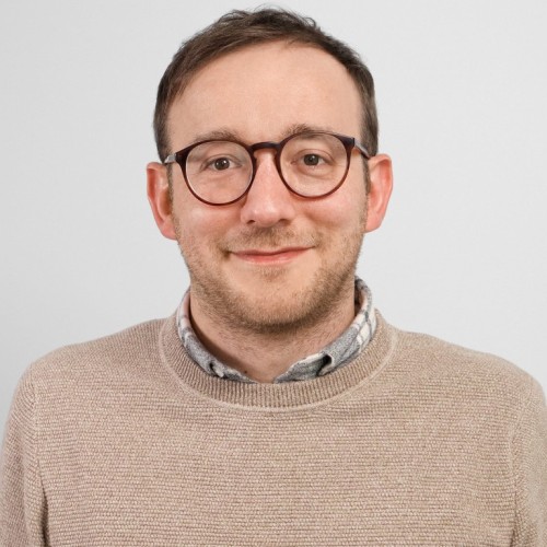 Porträt von Dax Werner. Er trägt eine runde Brille, einen beigefarbenen Strickpullover über einem karierten Hemd und blickt freundlich in die Kamera. Der Hintergrund ist neutral und hellgrau.