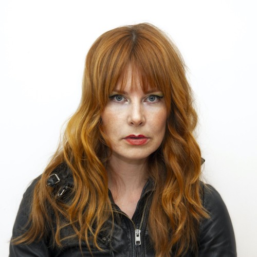 A headshot of a fair-skinned woman in her early thirties with long red hair, full lips, and a black leather jacket