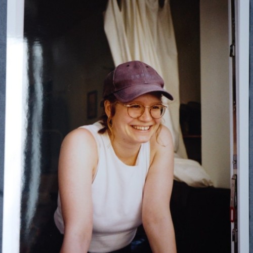 Ich stehe an einem offenen Fenster und stütze mich mit meinen Armen auf dem Fenstersims ab. Ich lächle und stehe dabei leicht schräg zum Fotografen. Ich schaue nicht in die Kamera, sondern lächele jemanden an, der neben der Kamera steht.