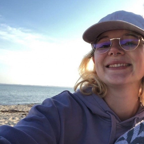 Eine Person mit Brille und Kappe im Vordergrund, Sand, Meer und Himmel im Hintergrund.
