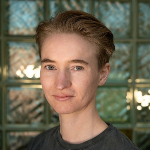 A portrait of Hari Klein in front of a blurred glass brick wall. 