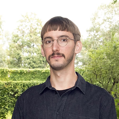 Ein Porträtfoto eines Mannes vor einer grünen, von Bäumen und Pflanzen geprägten Kulisse. Er trägt eine Brille, ein dunkles Hemd und hat einen kurzen Haarschnitt sowie einen Oberlippenbart.