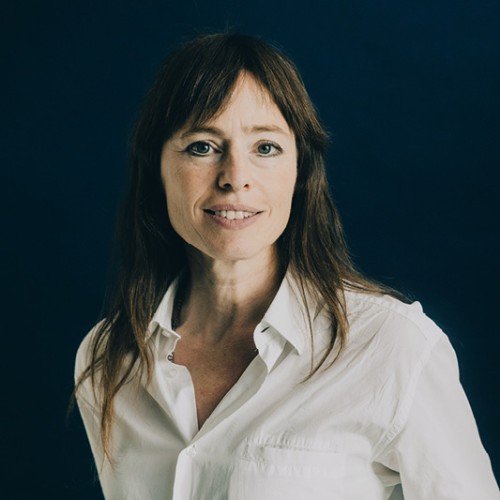 Hedwig Richter trägt lange glatte Haare und trägt eine Bluse. Sie schaut in die Kamera und lächelt. Dabei steht sie vor einem dunklen Hintergrund.