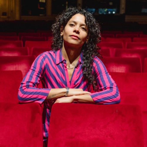 Auf dem Foto ist Emilia Roig in einem Theater- oder Kinosaal zwischen roten Vorstellungssesseln zu sehen. Sie trägt ein gestreiftes Oberteil und blickt direkt in die Kamera.
