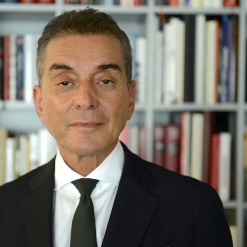 Auf dem Foto schaut Michel Friedman direkt in die Kamera. Er trägt einen schwarzen Anzug, ein weißes Hemd und eine schwarze Krawatte. Er steht vor einem Bücherregal.