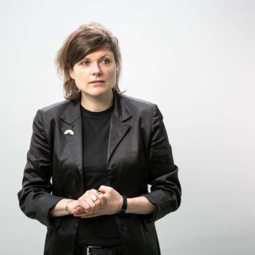 Portraitfoto von Tina Lorenz, eine nonbinary Person mit halblangen Haaren, die ein Tshirt und darüber ein Jackett trägt
