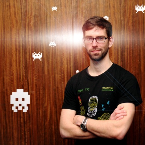 Ein viel zu junger Matthias mit kurzem Bart steht vor einer Holzwand mit weißen Spaceinvader-Pixel Figuren.