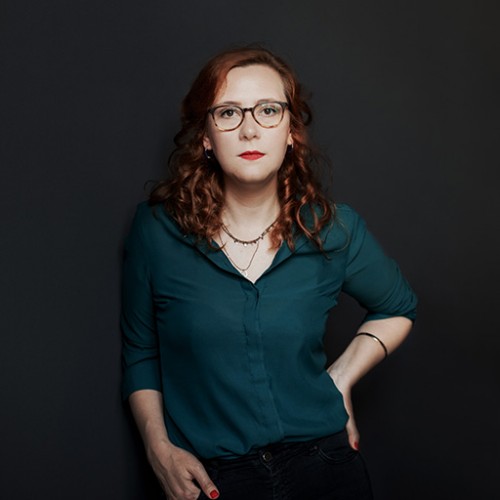  Anna ist sehr blass, trägt eine Brille und roten Lippenstift. Ihre Haare sind rot. Sie trägt eine petrolfarbene Bluse und hat eine Hand lässig an ihre Hüfte gestemmt.