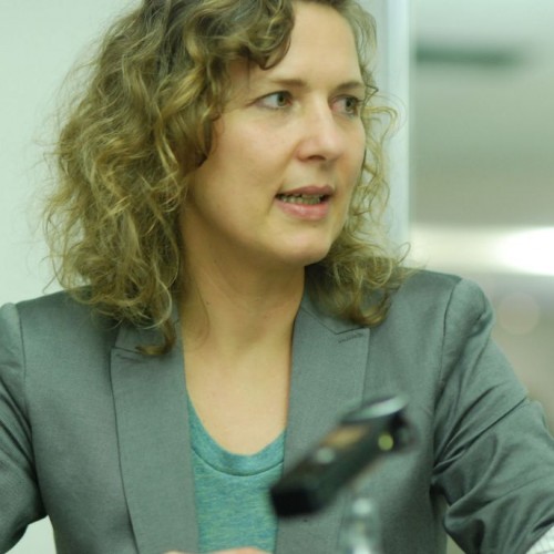Portrait von Ela Kagel mit grauem Jacket und grünem T-Shirt vor einem Mikrofon
